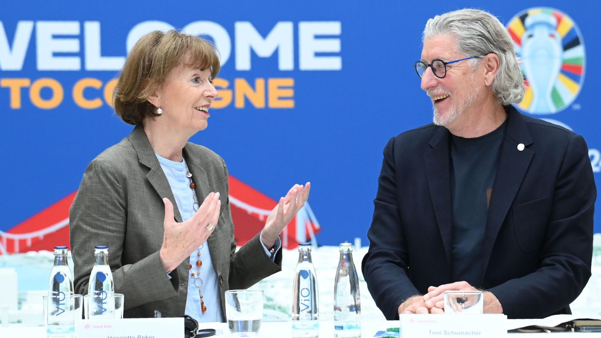 Zu sehen sind V.l. Henriette Reker und Toni Schumacher, die an einem Tisch stehen und lächeln.
