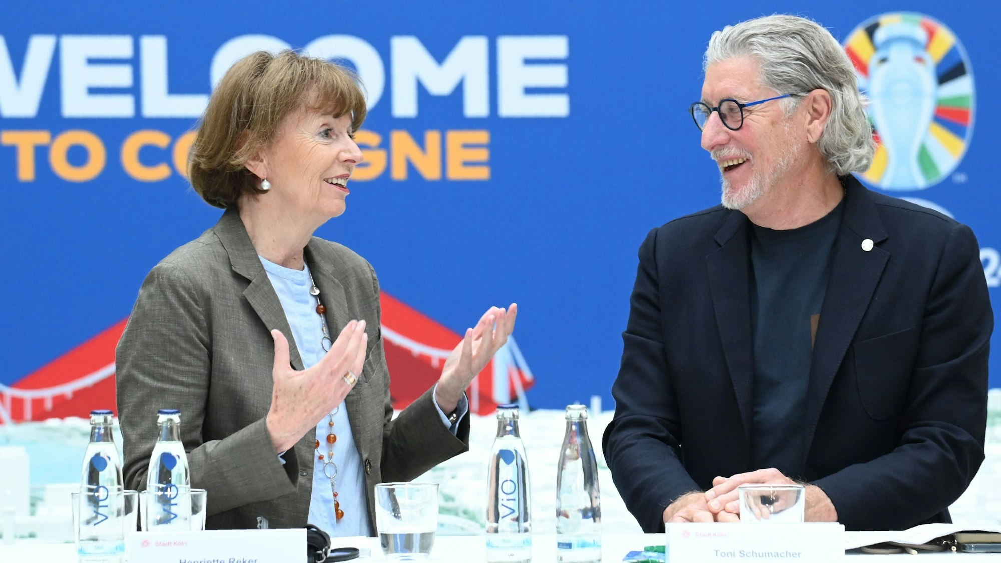 Zu sehen sind V.l. Henriette Reker und Toni Schumacher, die an einem Tisch stehen und lächeln.