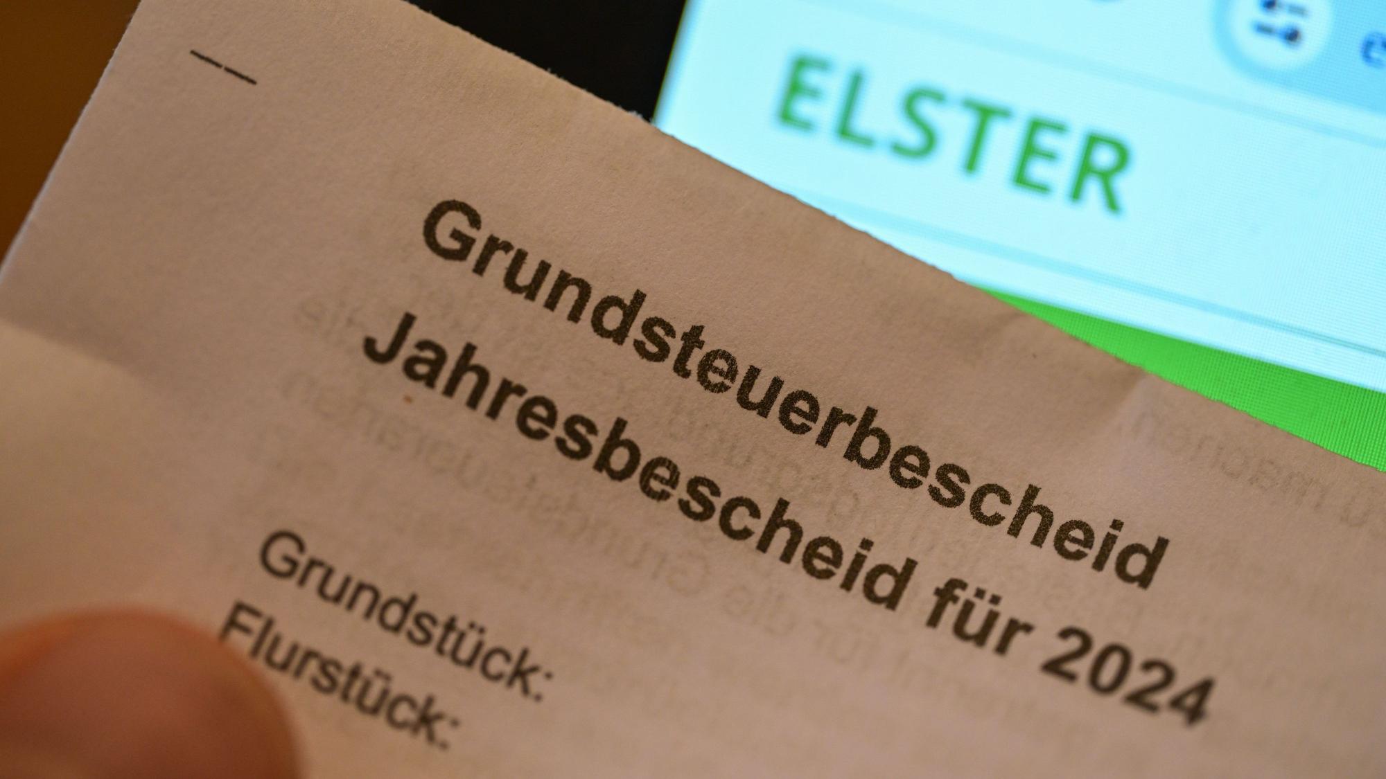 Das Bild zeigt einen Ausschnitt eines Grundsteuerbescheids. Im Hintergrund ist auf einem Bildschirm die Steuersoftware „Elster“ zu sehen.