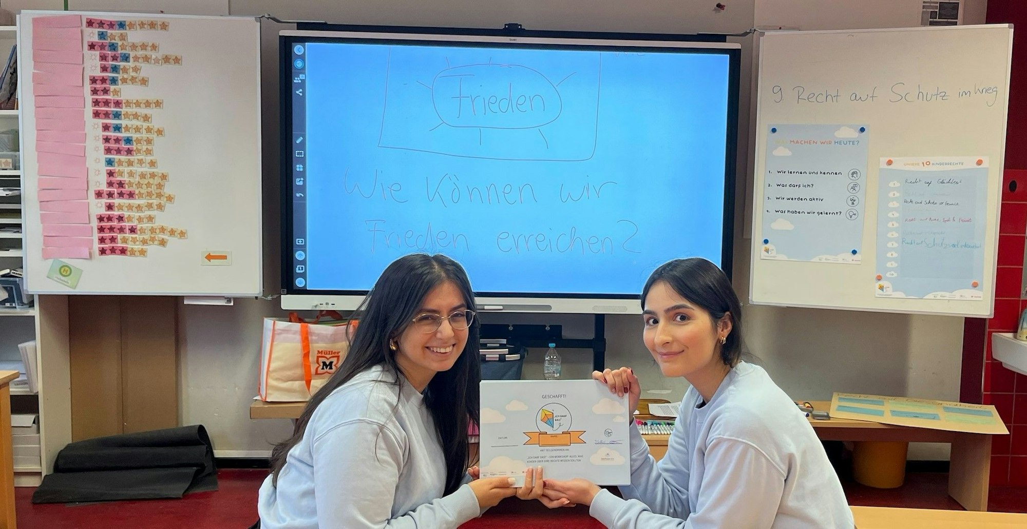 Elnara Valiyeva (links) und Jasmin Saffari-Esfahani haben den Kinderrechte-Workshop konzipiert, sie sitzen vor einer großen Schultafeln und halten das Logo des Workshops in der Hand.