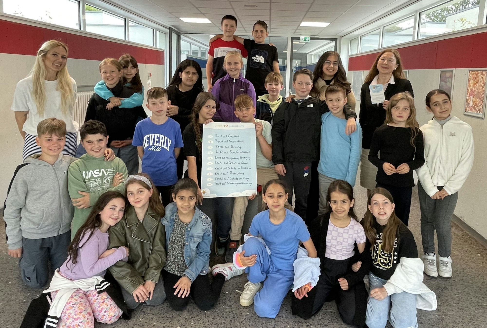 Schülerinnen und Schüler der vierten Klassen haben an der Gemeinschaftsgrundschule Köln-Flittard einen Kinderrechte-Workshop absolviert. Sie sitzen im Schulflur und halten ein Plakat mit den wichtigsten zehn Kinderrechten in die Höhe.