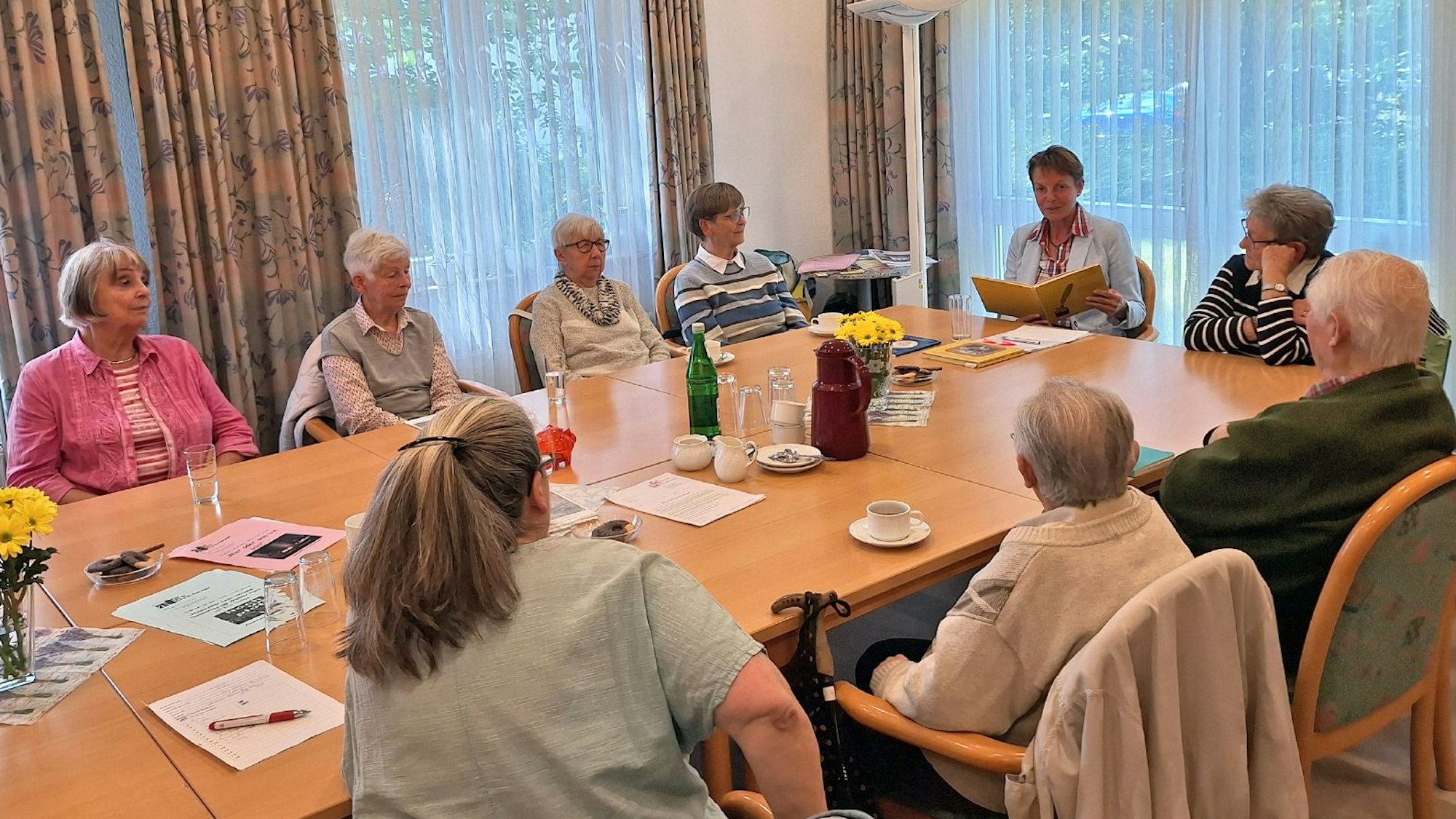 Eine Gruppe Senioren sitzt um einen Tisch herum, am Kopfende sitzt Iris Trespe mit einem aufgeschlagenen Buch.