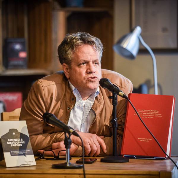 Marcus Bensmann sitzt an einem Tisch und spricht in ein Mikrofon. Auf dem Tisch sidn zwei Büchern auf Aufstellern. Bensmann trägt eine hellbraune Lederjacke und darunter ein weißes, gestreifes Hemd. Im Hintergrund weitere Bücher in einem Schrank.