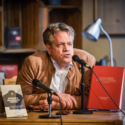 Marcus Bensmann sitzt an einem Tisch und spricht in ein Mikrofon. Auf dem Tisch sidn zwei Büchern auf Aufstellern. Bensmann trägt eine hellbraune Lederjacke und darunter ein weißes, gestreifes Hemd. Im Hintergrund weitere Bücher in einem Schrank.