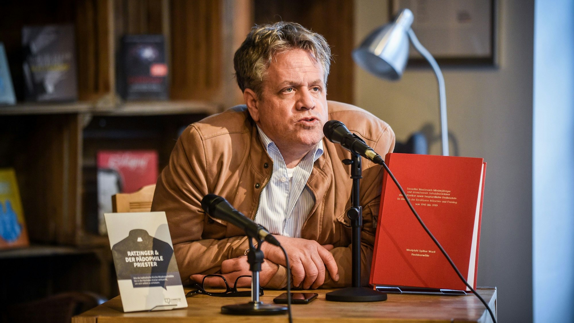 Marcus Bensmann sitzt an einem Tisch und spricht in ein Mikrofon. Auf dem Tisch sidn zwei Büchern auf Aufstellern. Bensmann trägt eine hellbraune Lederjacke und darunter ein weißes, gestreifes Hemd. Im Hintergrund weitere Bücher in einem Schrank.