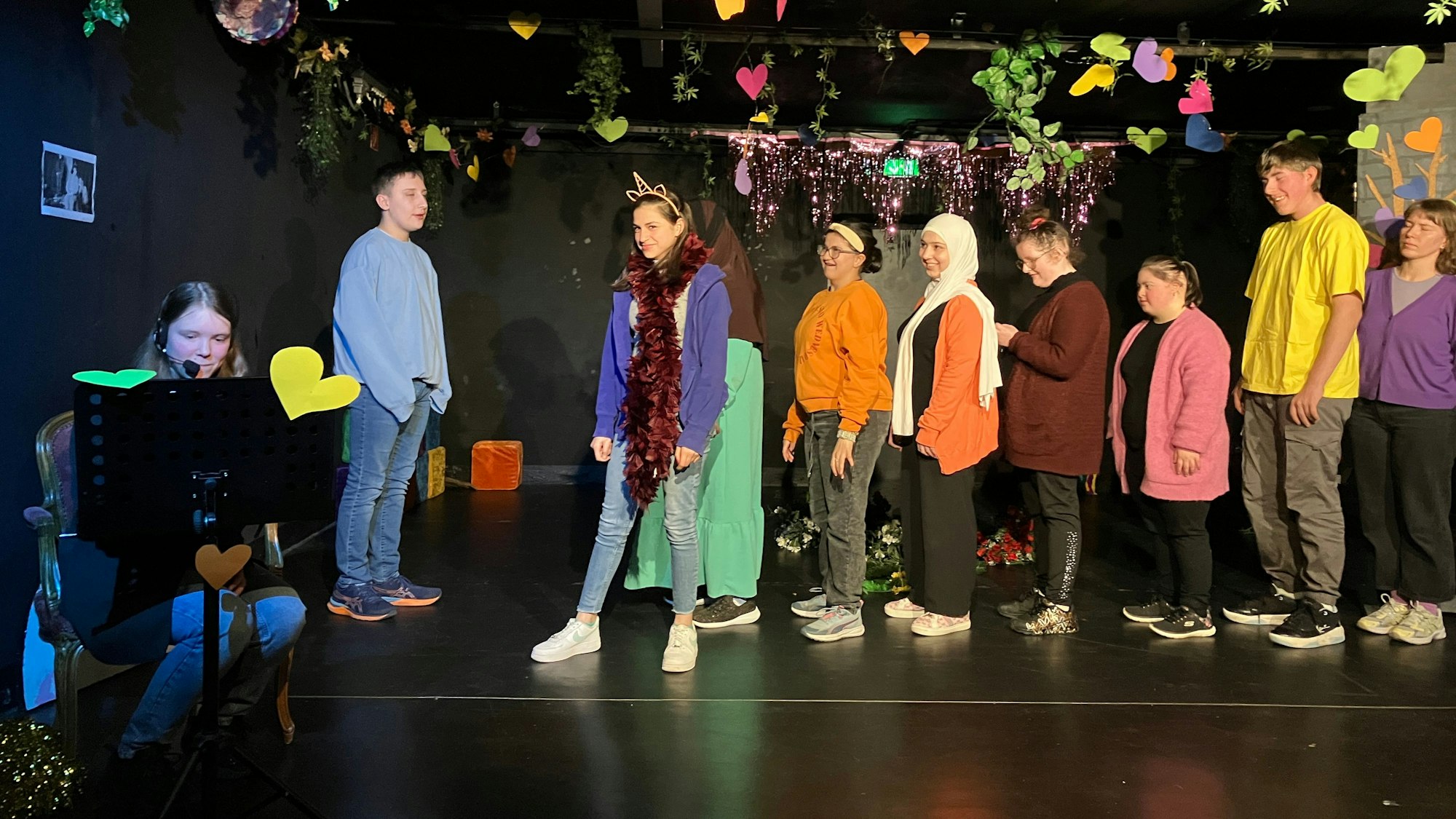 Auf der bunt geschmückten Bühne des Jungen Theaters sind acht Schauspielende zu sehen, die in einer Reihe hintereinander stehen, darunter das Nein-Horn, dessen Schauspielerin einen hornförmigen Haarreif und einen roten Glitzerschal trägt.