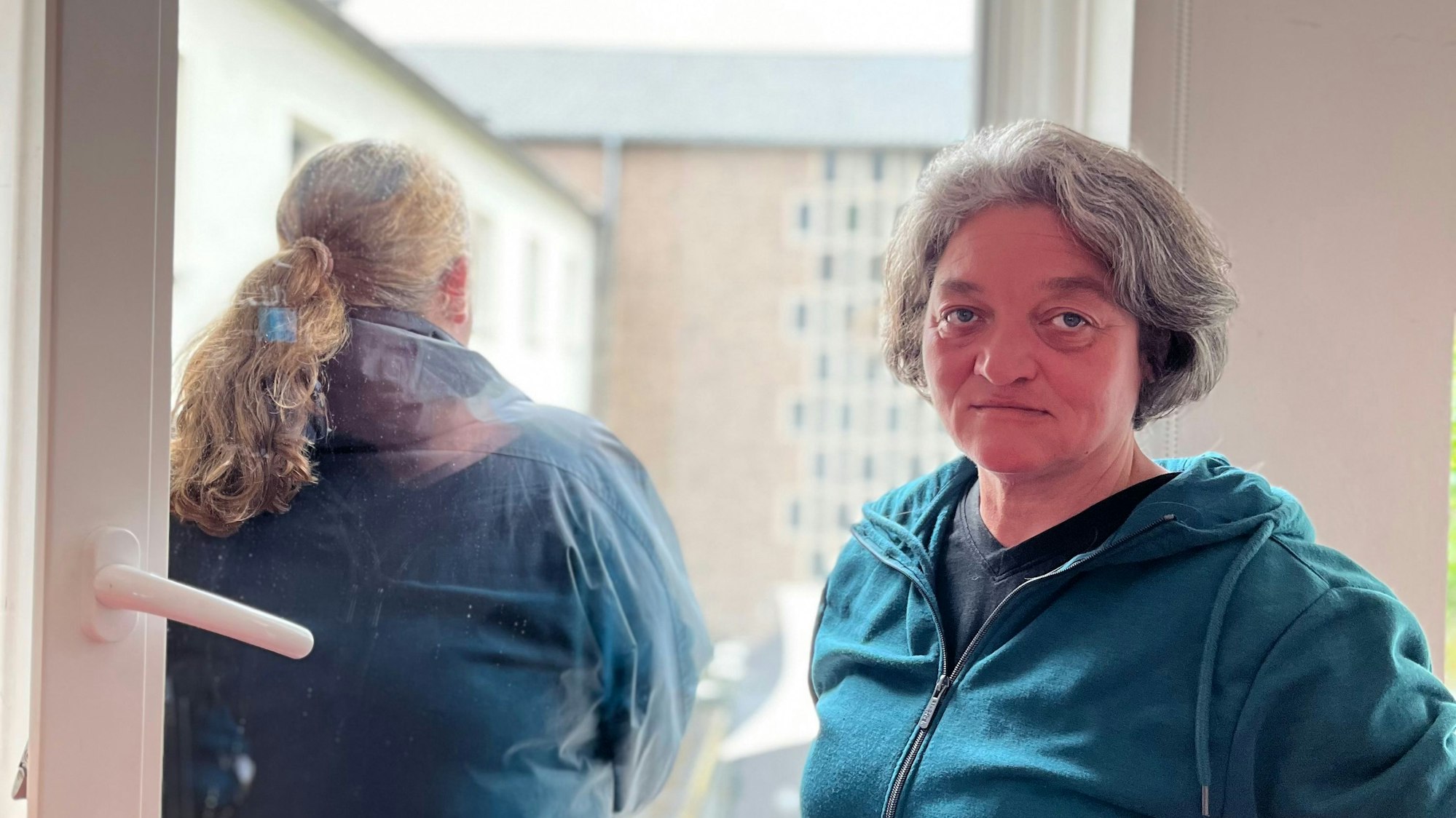 Eine Frau, Monika Wilke, gut in die Kamera, von einer zweiten ist eine Rückenansicht zu sehen, sie schaut aus dem Fenster.