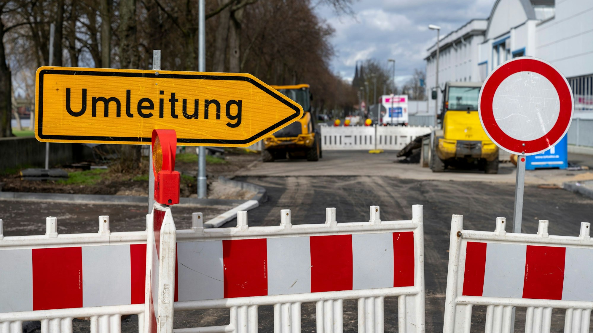 Auf dem Foto ist eine Baustelle am Rheinufer zu sehen. Ein orangenes Schild deutet auf die Umleitung des Verkehrs hin. Im Hintergrund ist der Dom zu sehen.