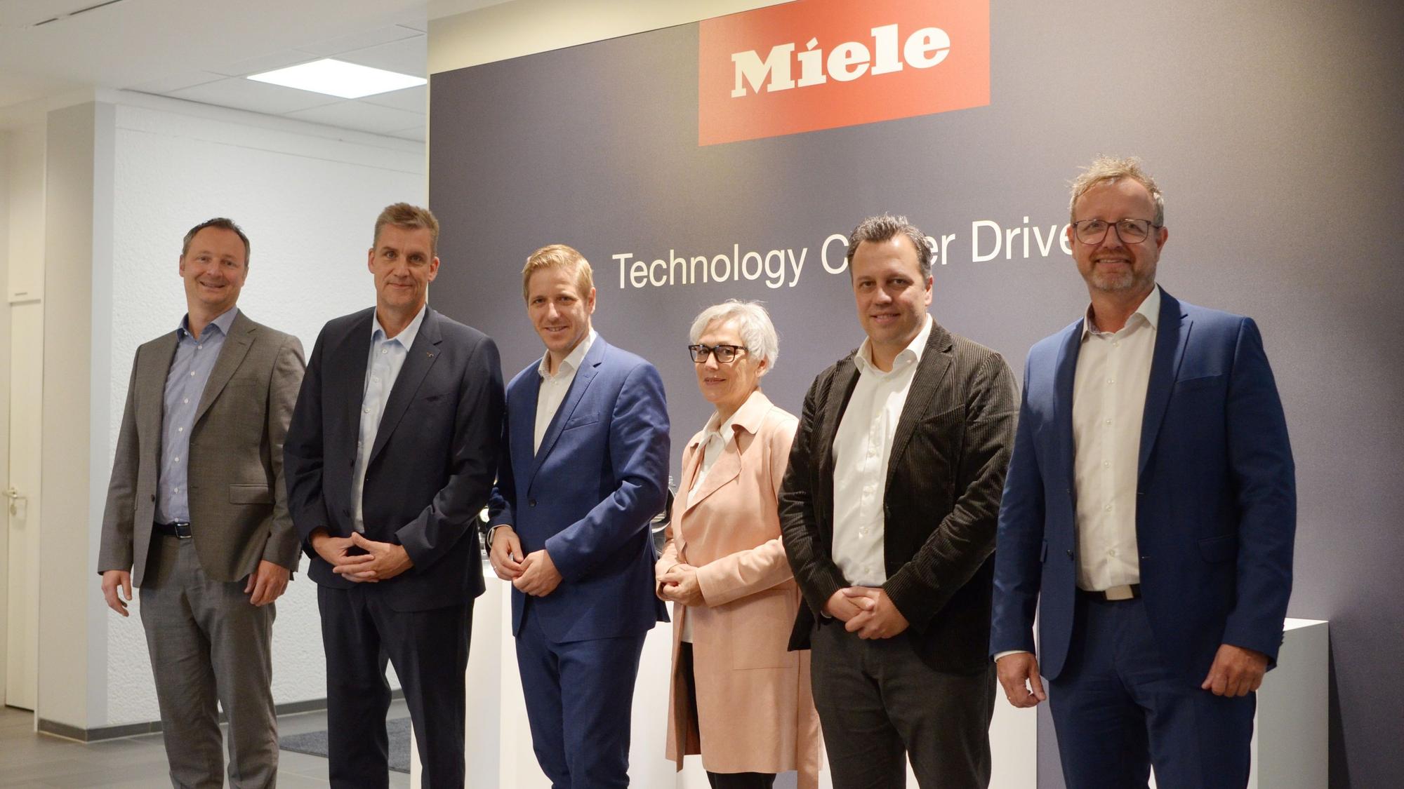 Gastgeber und Besucher stehen nebeneinander vor einer Wand mit dem Miele-Schriftzug.