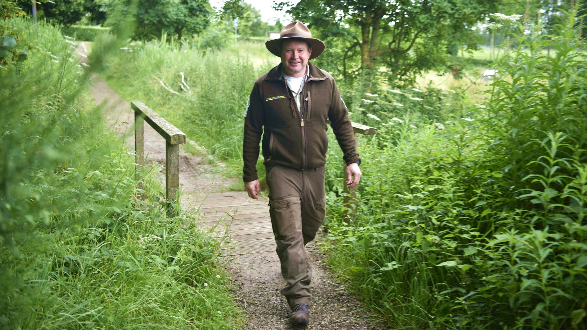 Nationalpark-Ranger Arno Koch läuft über eine kleine Brücke, rundherum sind Wiesen, Bäume, Brennnesseln und Blumen. Er trägt den typischen Ranger-Hut, eine braune Fleece-Jacke und eine hellbraune Hose.