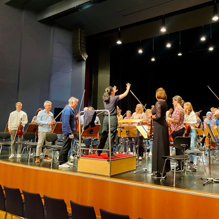 Bar Avni steht auf einem Podest vor dem Orchester. Auch die Musikerinnen und Musiker haben sich erhoben.