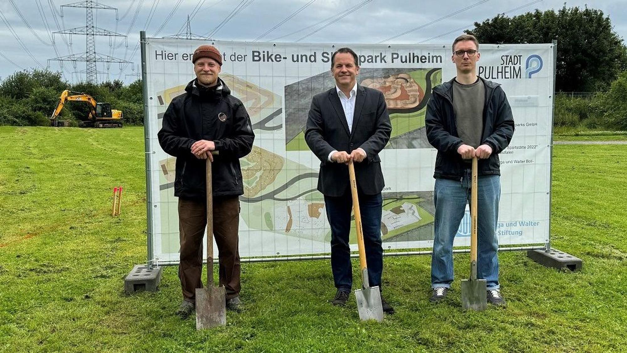 Drei Männer stehen vor einem Transparent. Jeder hat einen Spaten in der Hand.
