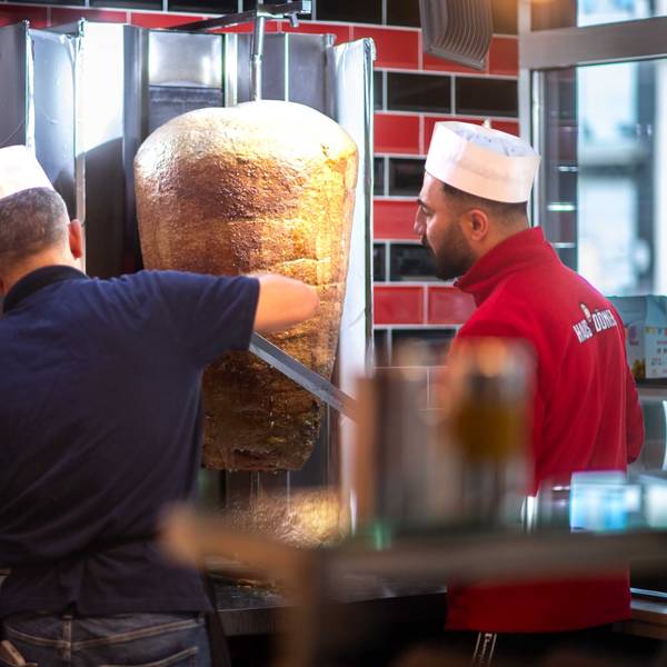 Das Bild zeigt zwei Mitarbeiter, die von zwei Döner-Spießen Fleisch abschneiden.