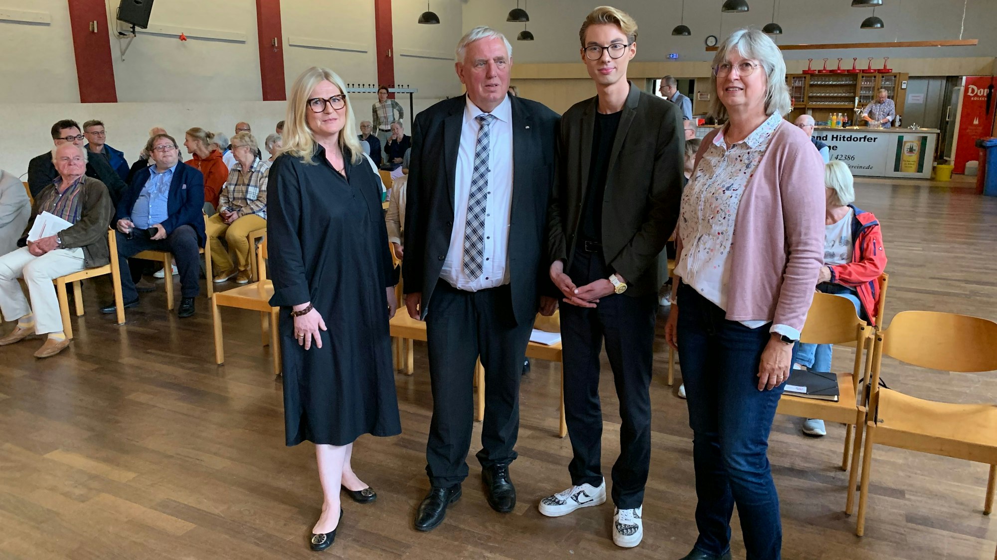 Zwei Männer und zwei Frauen stehen in einer Halle vor Publikum.