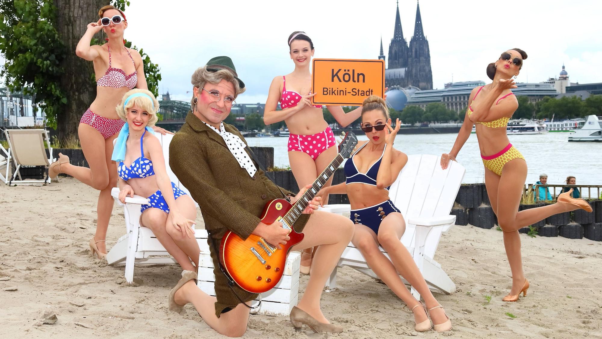 Frauen posieren im Bikini am Rheinufer, davor hockt ein Mann im Oma-Kostüm mit Gitarre.