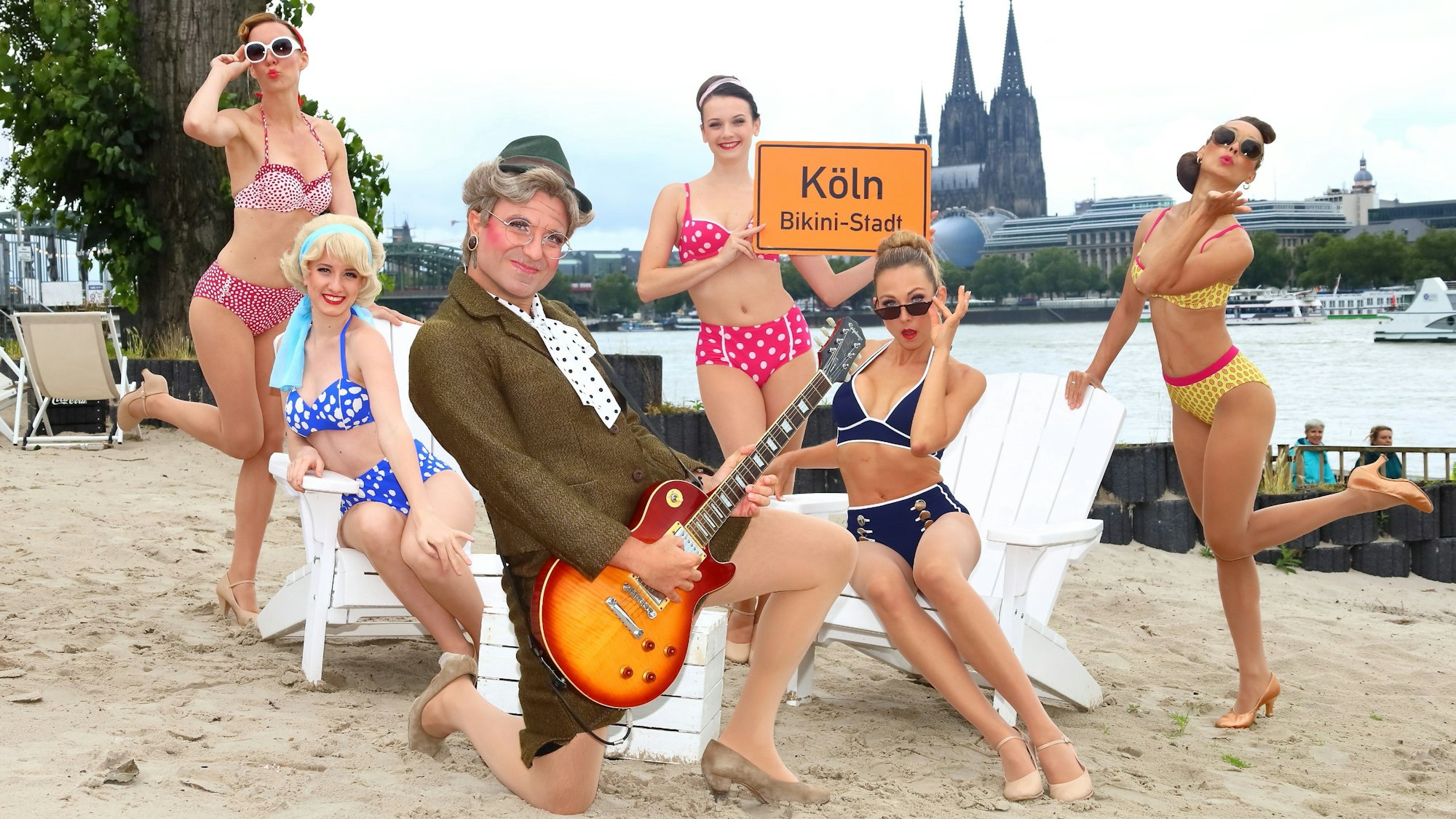 Frauen posieren im Bikini am Rheinufer, davor hockt ein Mann im Oma-Kostüm mit Gitarre.