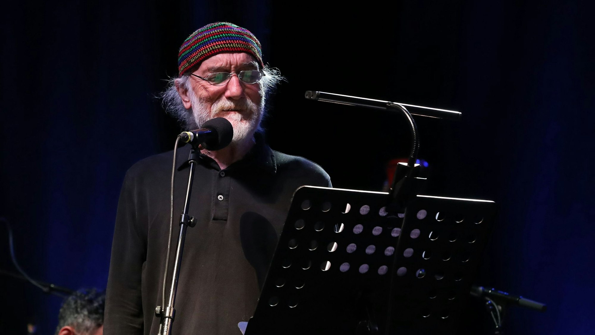 Fuat Saka steht auf einer Bühne, er hat eine gestreifte Wollmütze auf und trägt ein schwarzes Hemd. Er hat einen weißen Bart und trägt eine Brille. Er steht vor einem Mikrofon und einem Notenpult.