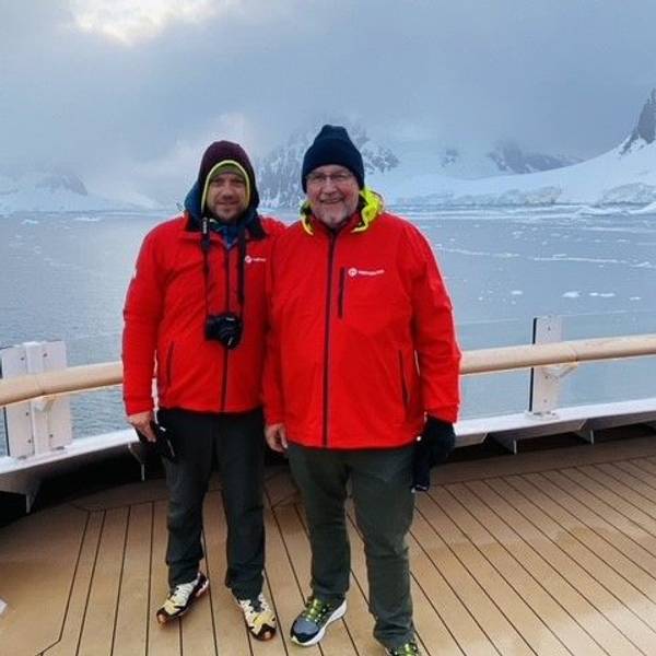 Christopher und Erich Koch mit roten Jacken stehen auf einem Schiffsdeck, im Hintergrund sind eisbedeckte Berge zu sehen.