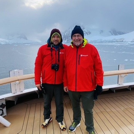 Christopher und Erich Koch mit roten Jacken stehen auf einem Schiffsdeck, im Hintergrund sind eisbedeckte Berge zu sehen.