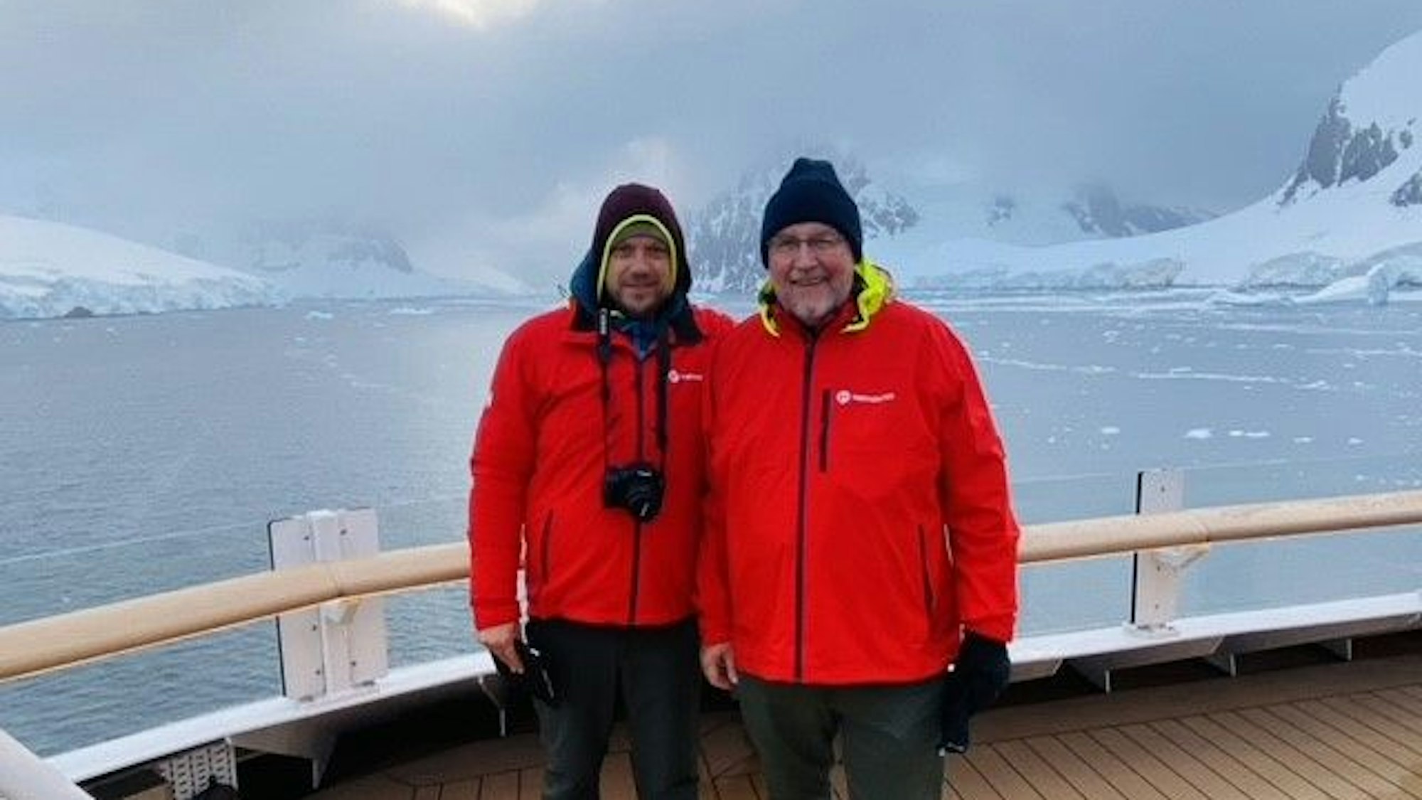 Christopher und Erich Koch mit roten Jacken stehen auf einem Schiffsdeck, im Hintergrund sind eisbedeckte Berge zu sehen.