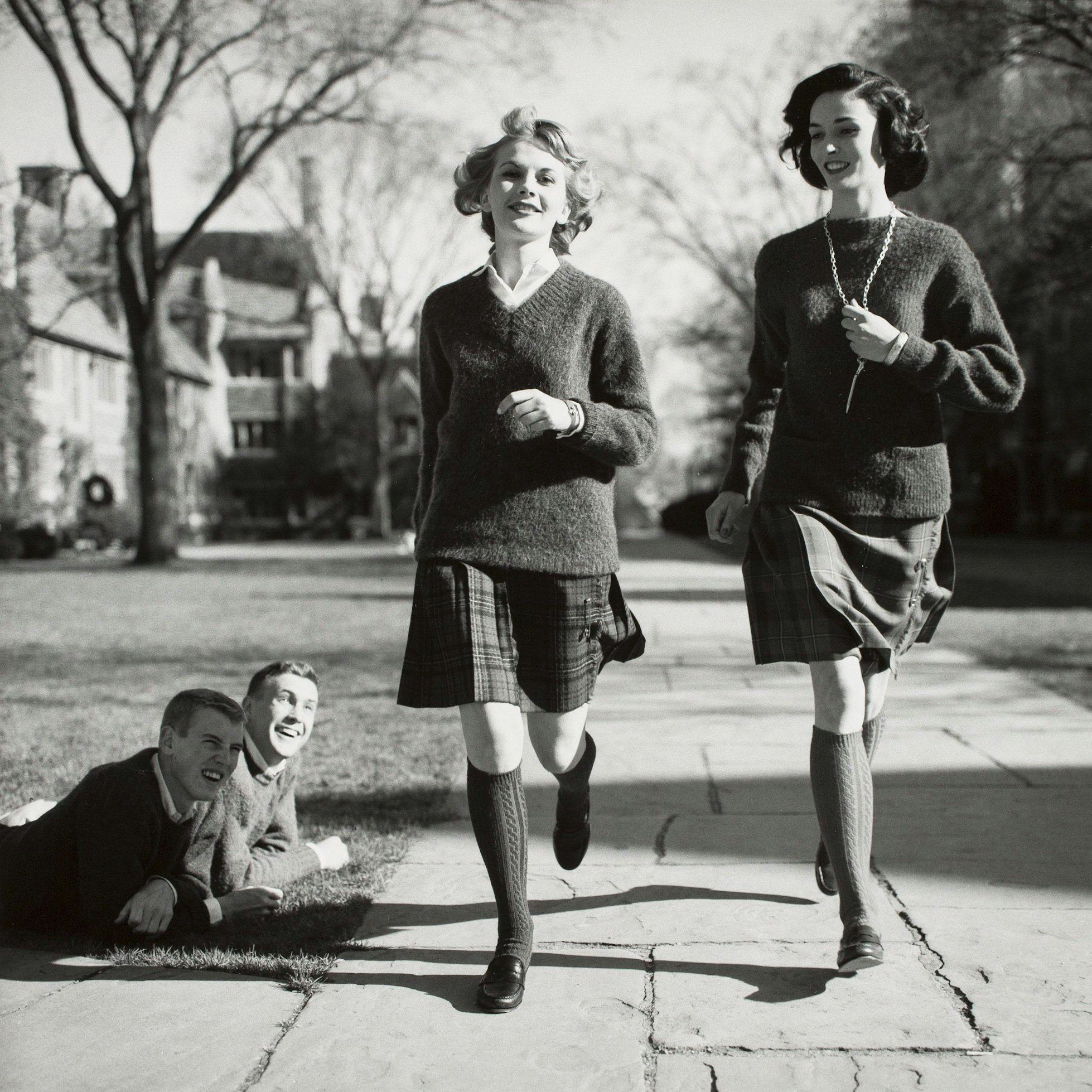 Die Schwarz-weiß-Aufnahme von 1958 zeigt zwei junge Frauen, die Kniestrümpfe, Röcke und Pullover tragen und an zwei Männern vorbeigehen, die auf einer Rasenfläche liegen und ihnen hinterherschauen.
