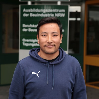 Der junge Mann ist im Halbporträt zu sehen. Er trägt einen Kapuzenpullover und lächelt in die Kamera.