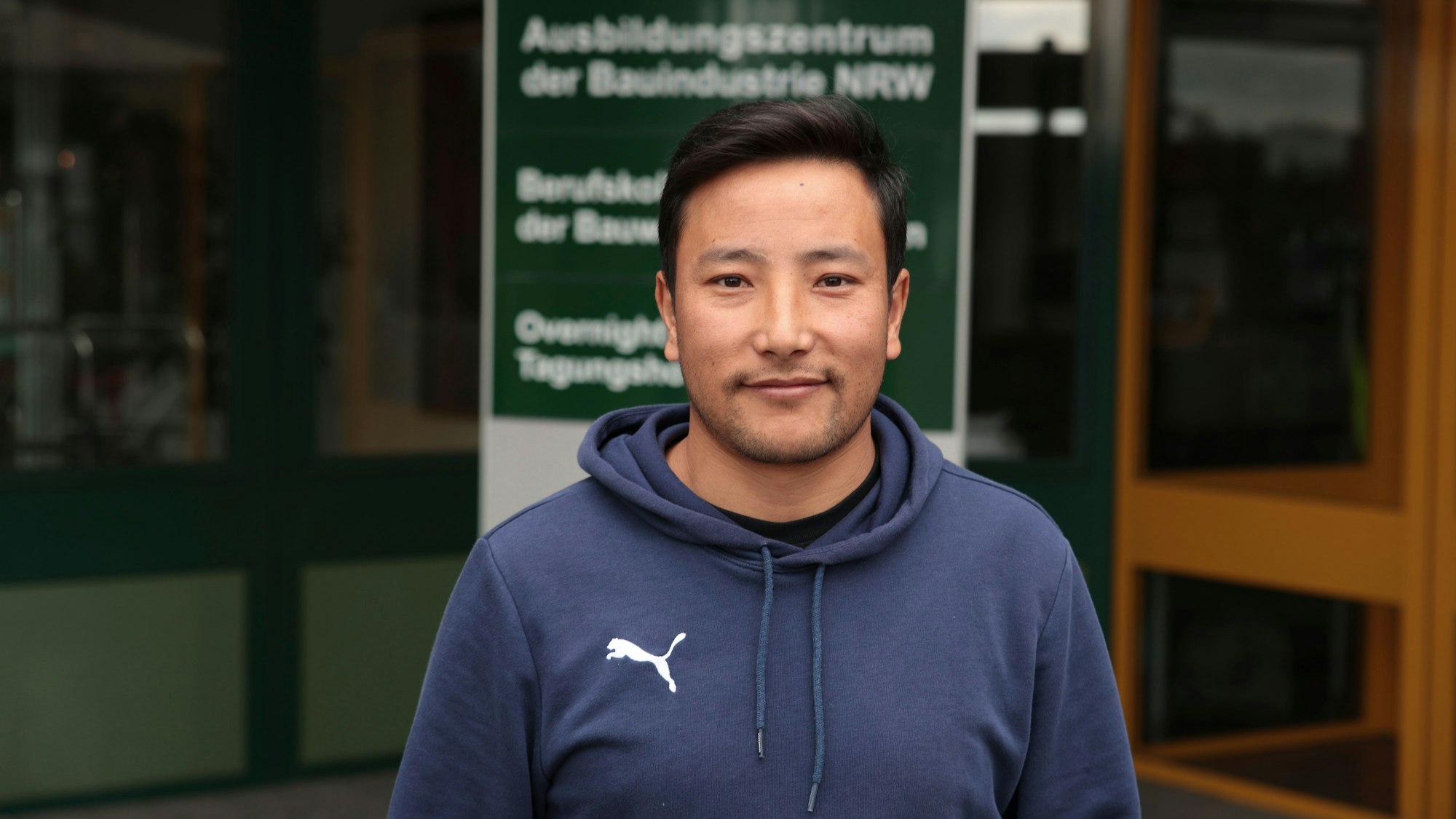 Der junge Mann ist im Halbporträt zu sehen. Er trägt einen Kapuzenpullover und lächelt in die Kamera.