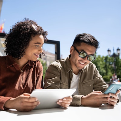 Ein Mann und eine Frau sitzen bei Sonne auf einer Terrasse und schauen auf ein Tablet sowie auf ein Smartphone