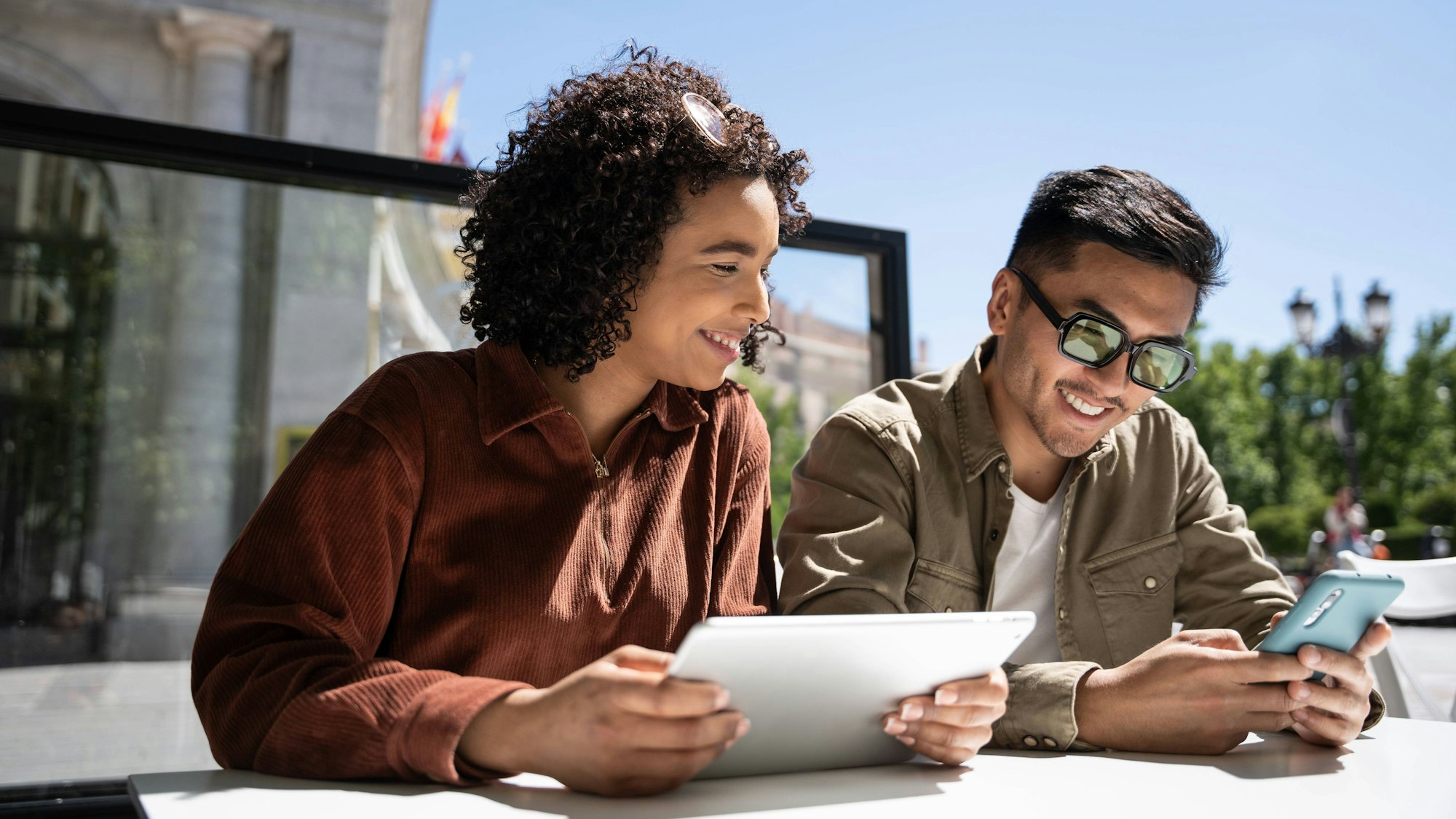Ein Mann und eine Frau sitzen bei Sonne auf einer Terrasse und schauen auf ein Tablet sowie auf ein Smartphone