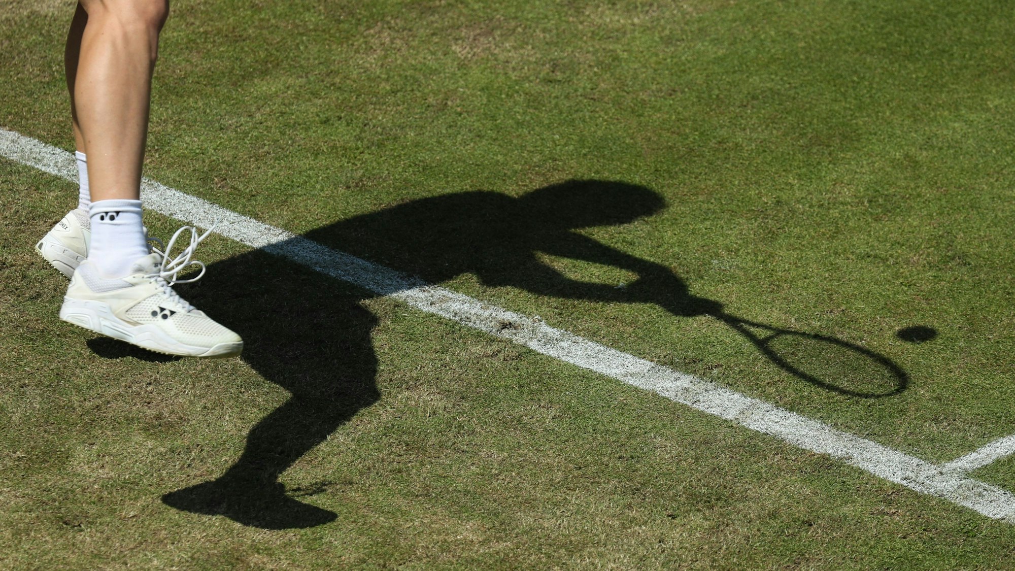 Ein Herrenbein ist oben rechts im Bild zu sehen. Tennisschuhe und Tennissocken werden getragen. Ein Schatten ist zentraler Inhalt des Bildes, man sieht einen Mann einen Tennisball mit einem Tennisschläger schlagen.