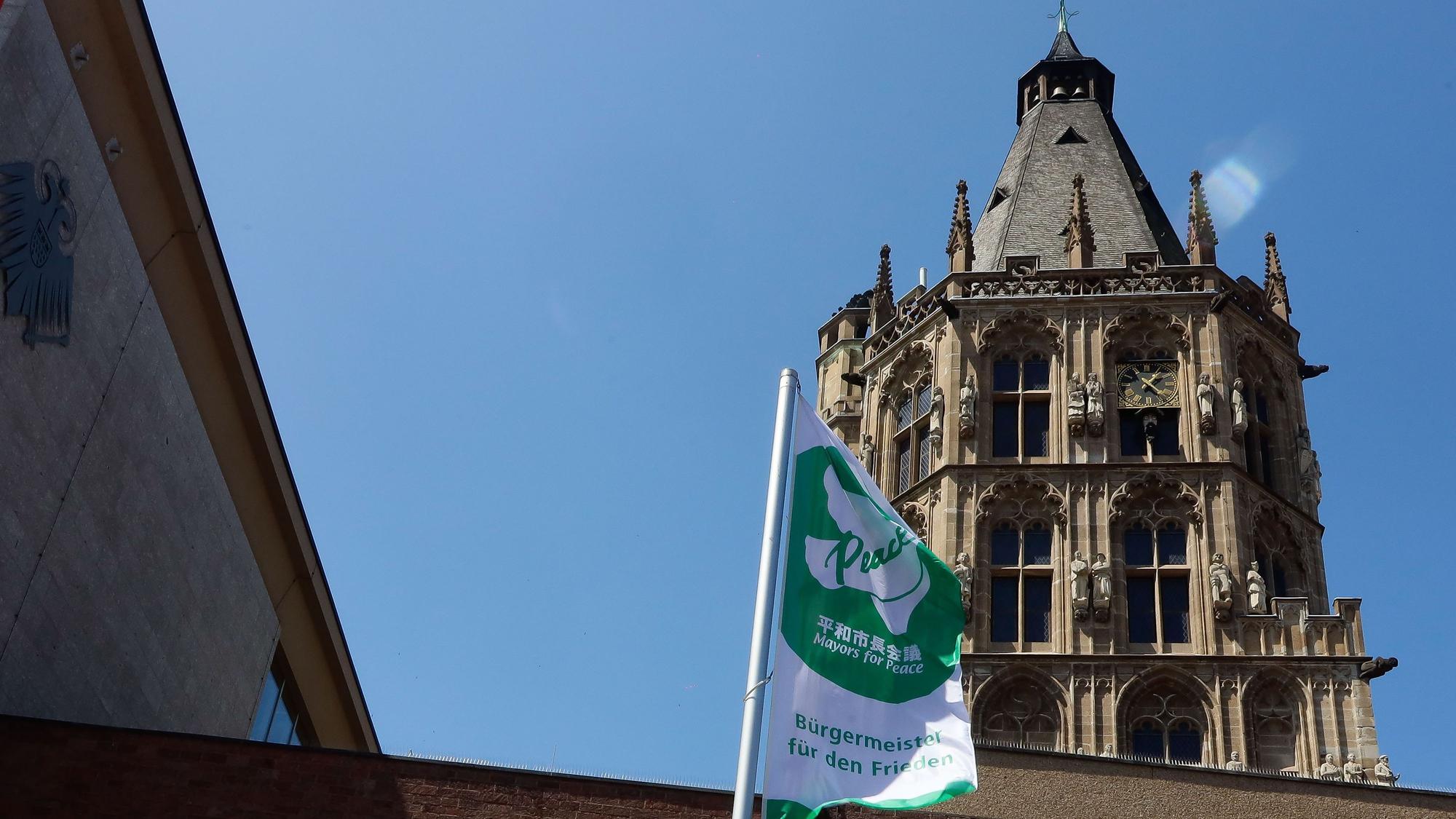 Das Bild zeigt die weiß-grüne Flagge von „Mayors For Peace“ vor dem Rathausgebäude. Im Hintergrund ist der Rathausturm zu sehen.