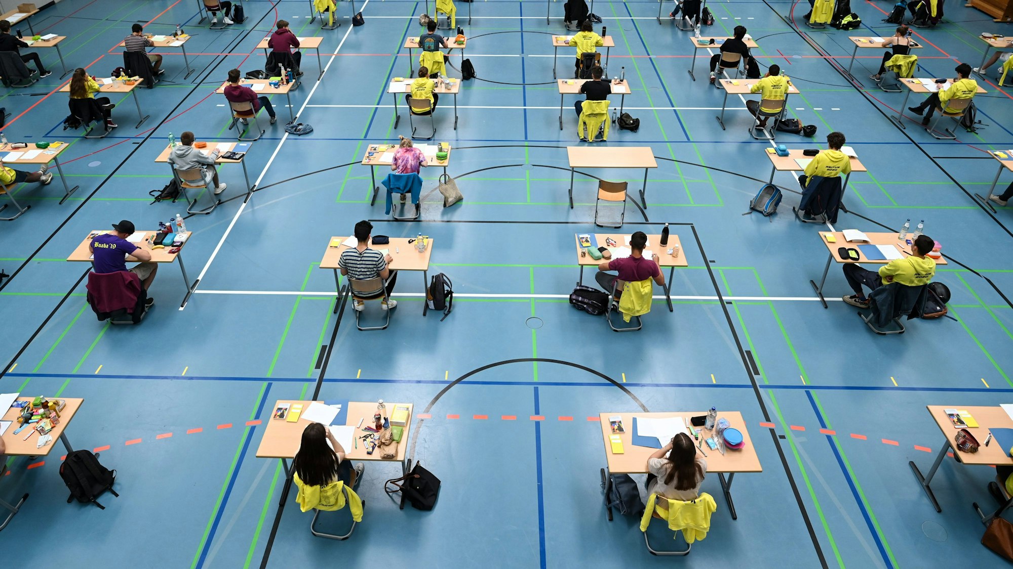 Schülerinnen und Schüler schreiben eine Prüfung in einer Turnhalle. Die Tischen sind mit viel Abstand und in langen Reihen angeordnet.