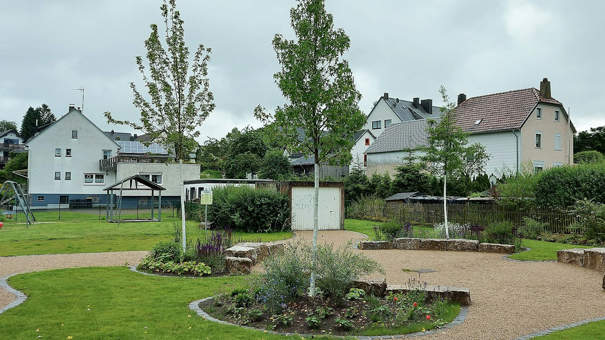 Das Bild zeigt einen Platz mit Bäumen, Bänken und Blumenbeeten.