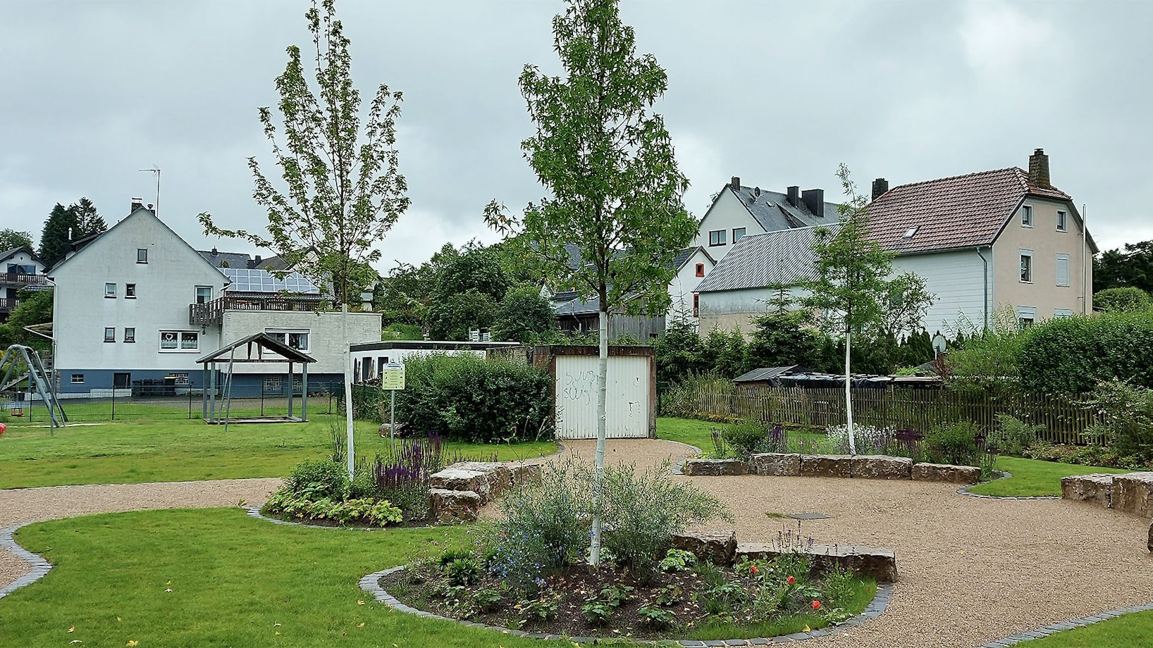 Das Bild zeigt einen Platz mit Bäumen, Bänken und Blumenbeeten.