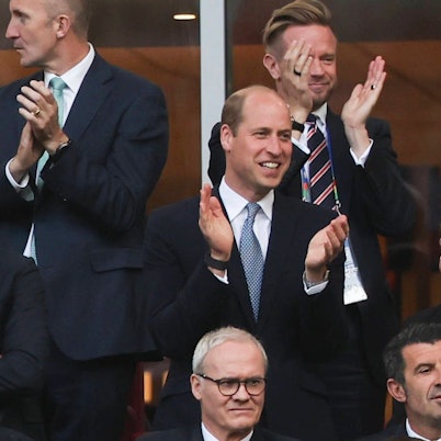 Prinz William klatscht grinsend in die Hände während er auf einer Tribüne steht.