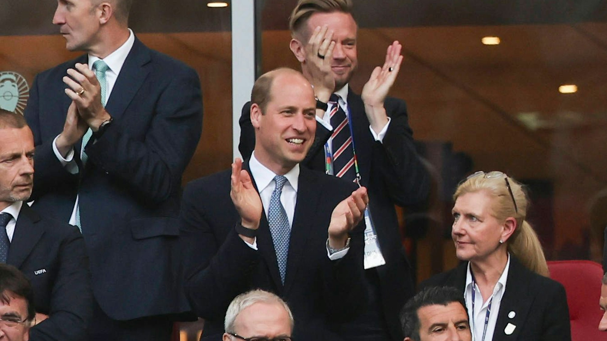 Prinz William klatscht grinsend in die Hände während er auf einer Tribüne steht.