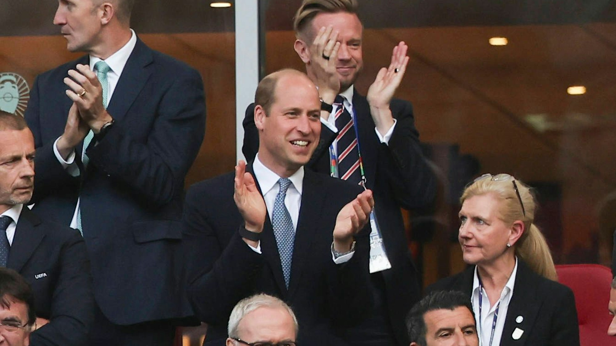 Prinz William klatscht grinsend in die Hände während er auf einer Tribüne steht.