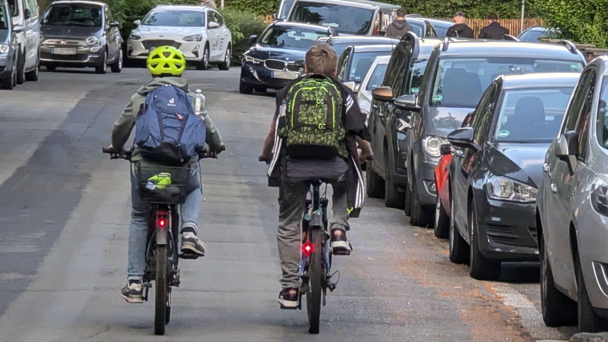 Zwei Kinder mit Rucksäcken auf dem Rücken fahren nebeneinander mit Fahrrädern über die Bonnstraße.