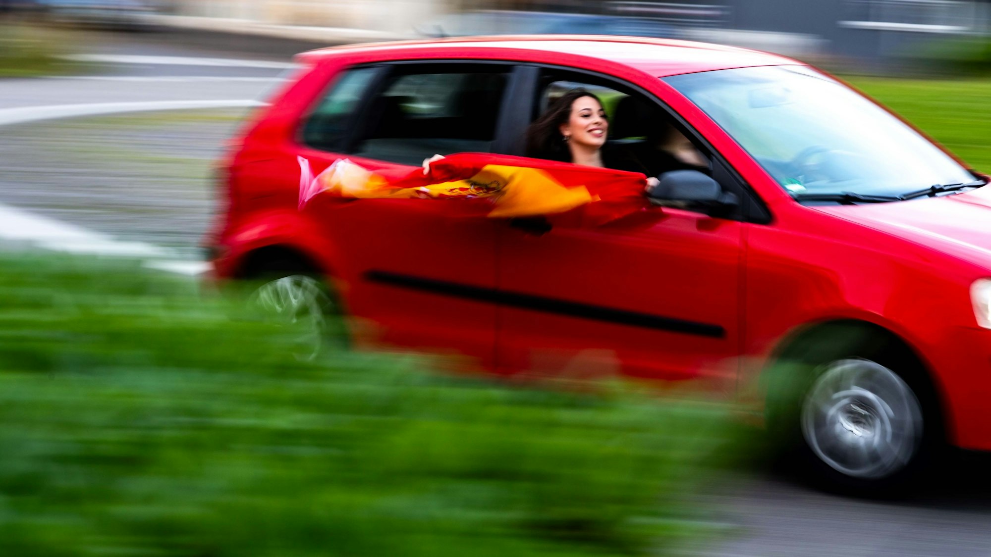 Das Bild zeigt ein rotes Auto, aus dem eine junge Frau eine spanische Flagge hält.