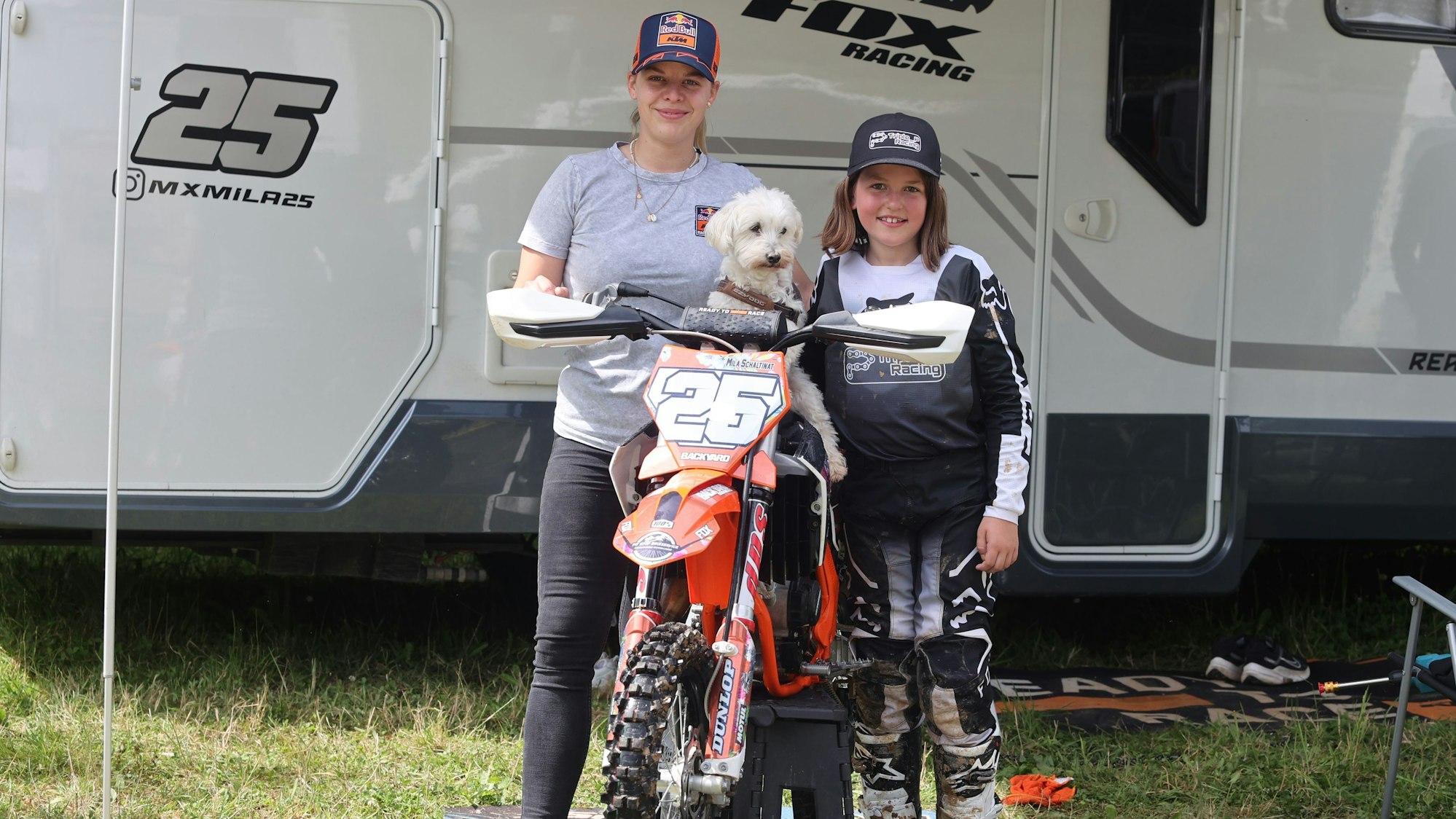 Foto einer jungen Motocross-Fahrerin mit ihrer Maschine, ihrer Mutter und einem Hund.