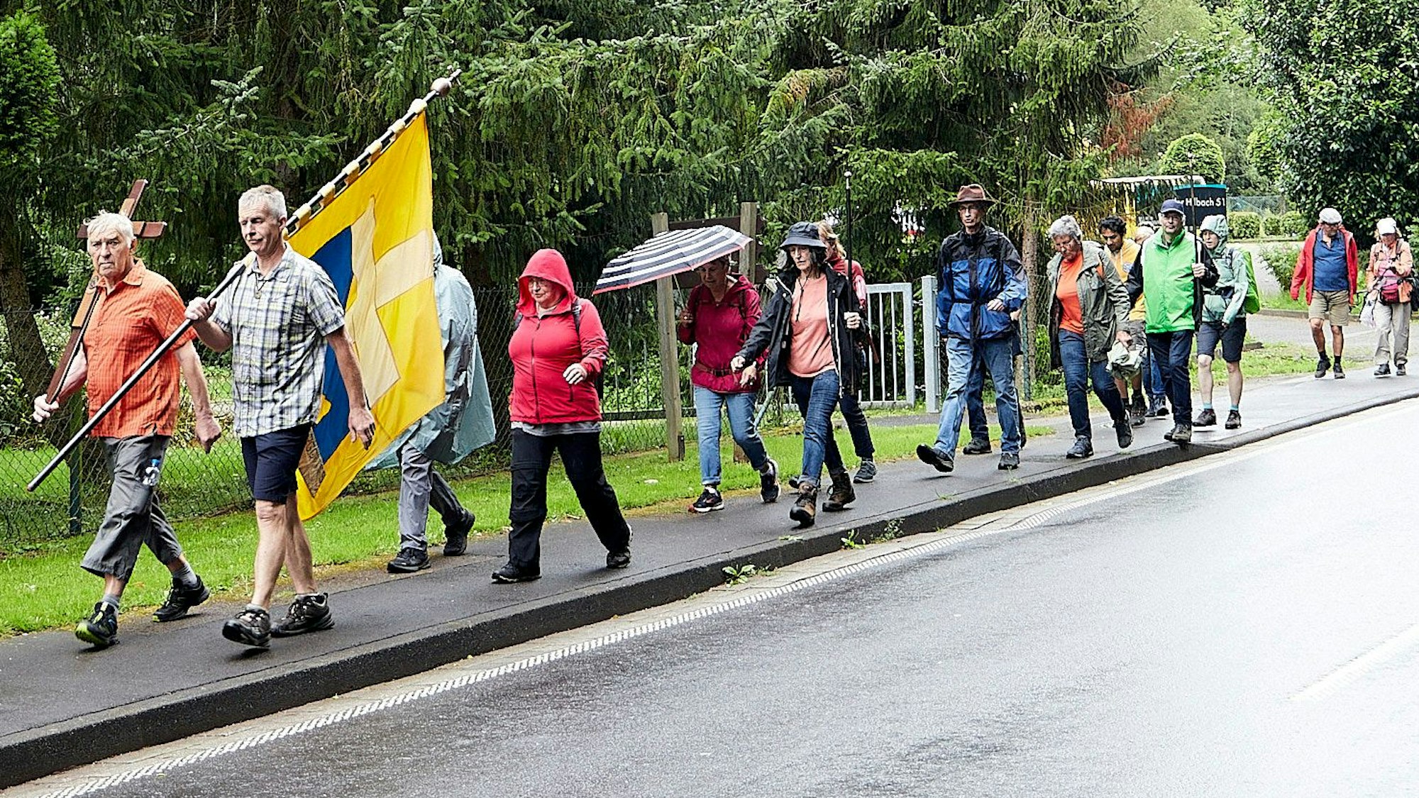 Eine Gruppe Pilger aus Niederzier geht im Regen auf einem Gehweg. Die beiden Männer, die vorangehen, tragen eine Fahne und ein Kreuz.