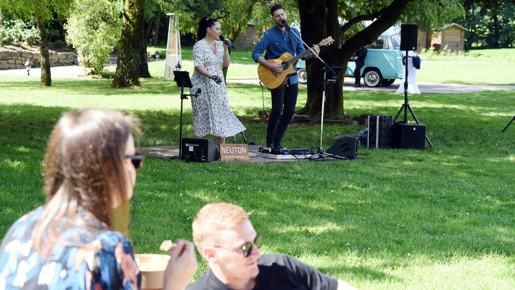 Zwei Musiker spielen und singen auf einer grünen Wiese, auf der Menschen picknicken.