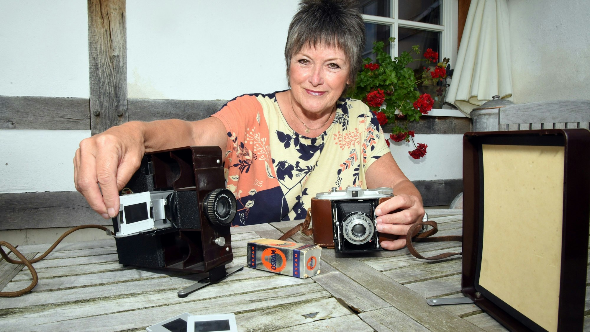 Eine Frau sitzt an einem Gartentisch und hält eine Hand über einen Diaprojektor. Auf dem Tisch liegen außerdem Dias, eine Leinwand und eine alte Fotokamera.