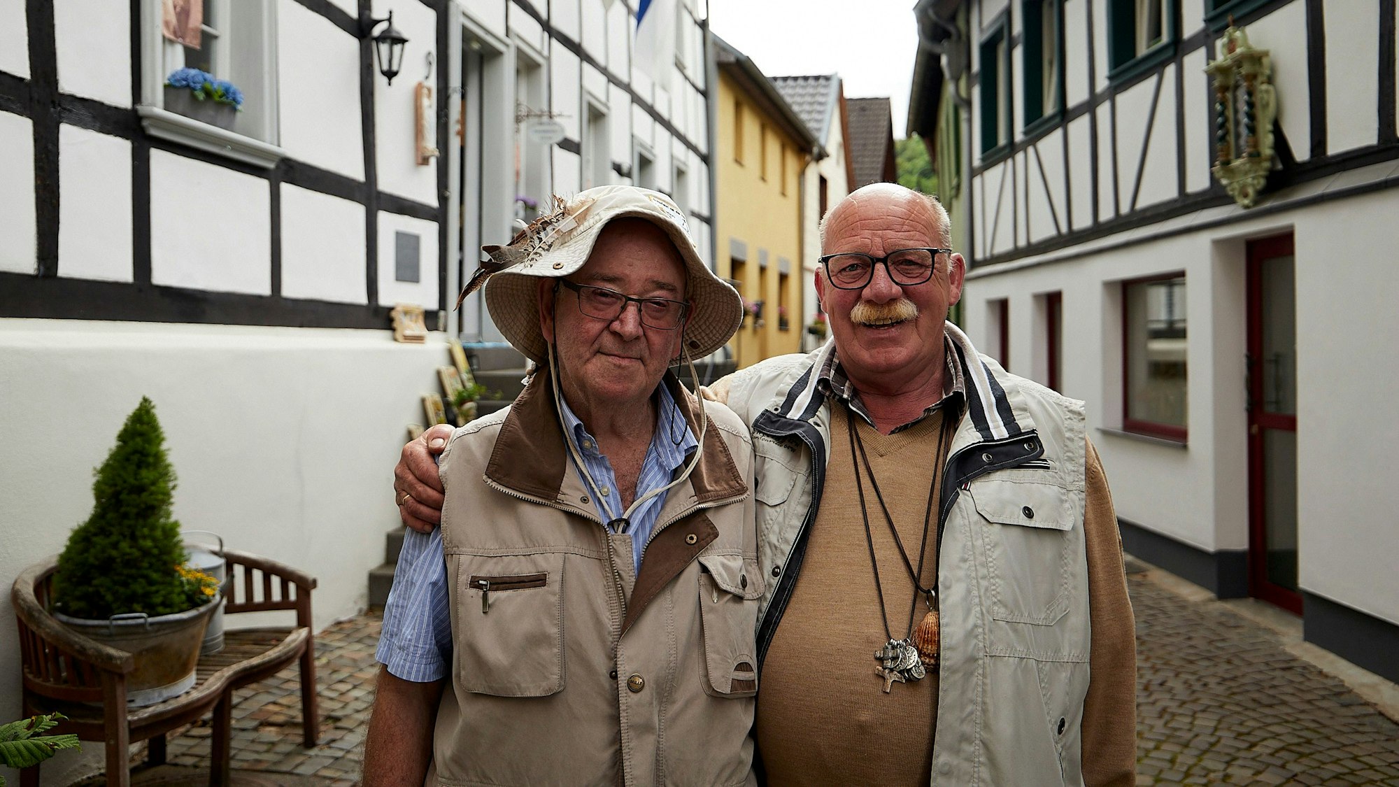 Zwei Männer, Pilgervater Helmut Heuer und Pilger Alois Neumann, stehen Arm in Arm in einer Gasse in Heimbach, die von Fachwerkhäusern gesäumt ist.