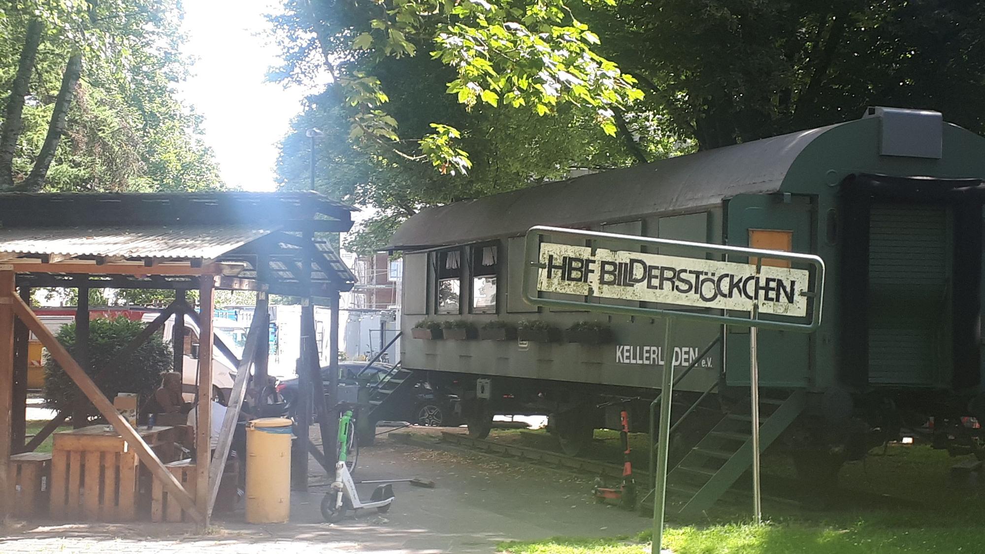 Links im Bild der überdachte Biergarten, rechts ein alter, dunkelgrüner Bahnwagon mit dem Schriftzug „Kellerladen“. Im Vordergrund steht ein Schild mit der Aufschrift „Hbf Bilderstöckchen“, im Hintergrund stehen Bäume.