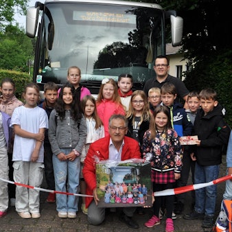 Grundschulkinder aus Golbach beim Abschiedsfoto mit Borislav Radovic. Sie stehen hinter einem Flatterband vor dem Schulbus, Radovic kniet bei den Kindern und hält eine Fotografie in Händen.