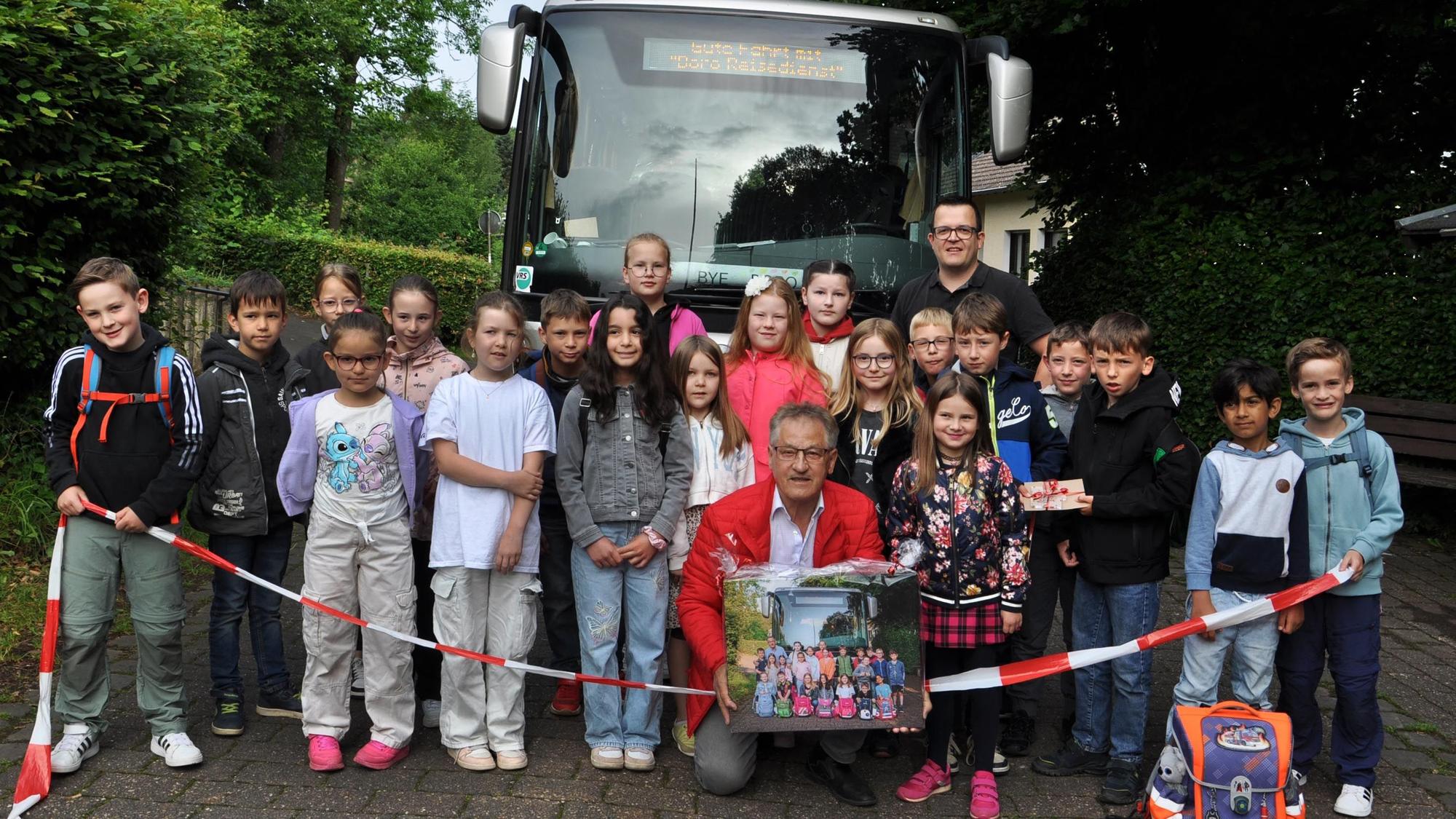 Grundschulkinder aus Golbach beim Abschiedsfoto mit Borislav Radovic. Sie stehen hinter einem Flatterband vor dem Schulbus, Radovic kniet bei den Kindern und hält eine Fotografie in Händen.