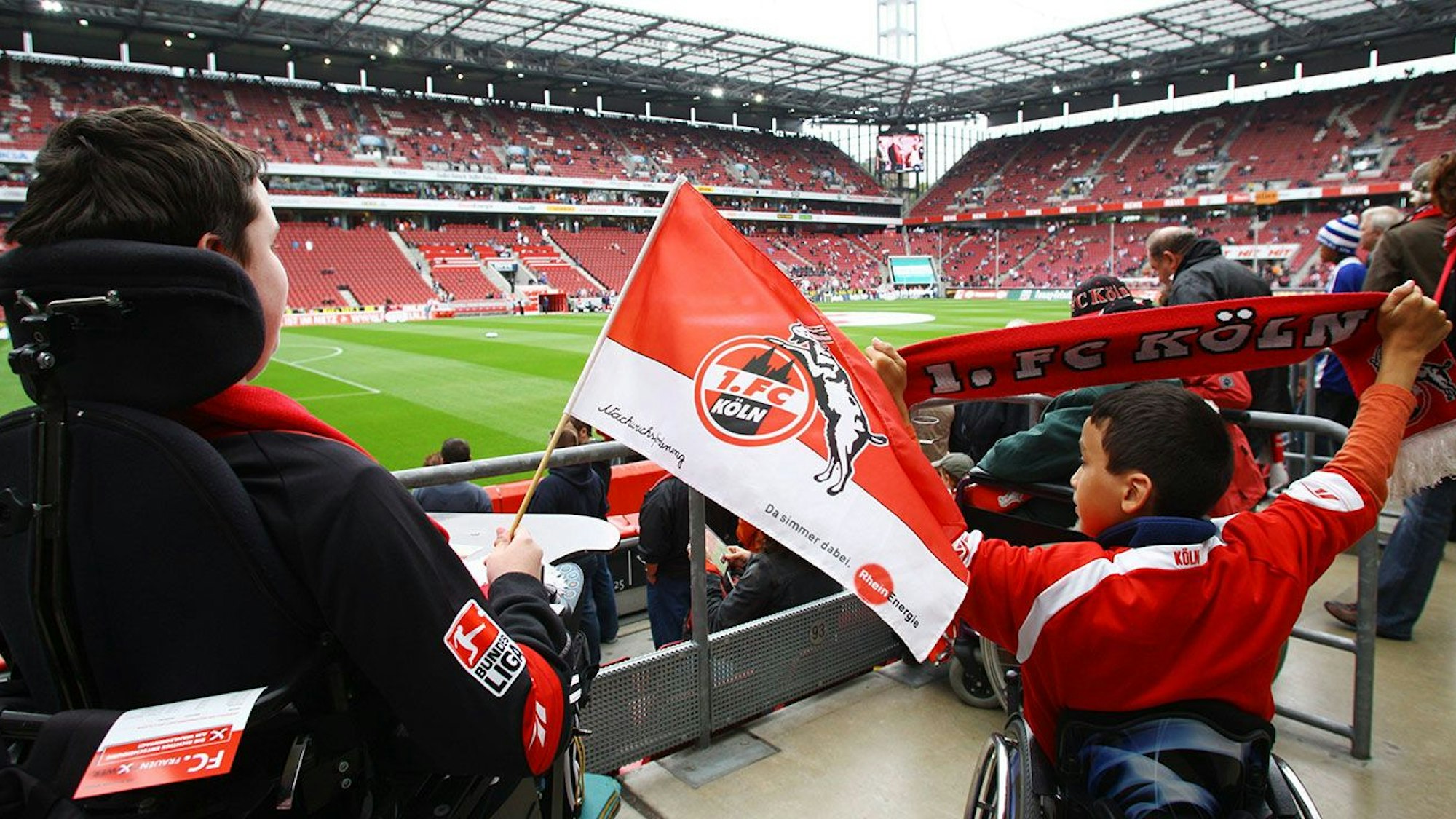 Ein Junge im Rollstuhl mit einer kleinen FC-Fahne in der Hand: 100 Rollstuhlfahrer und deren Begleiter können derzeit im Rheinenergie-Stadion bei einem Spiel des 1. FC Köln dabei sein.