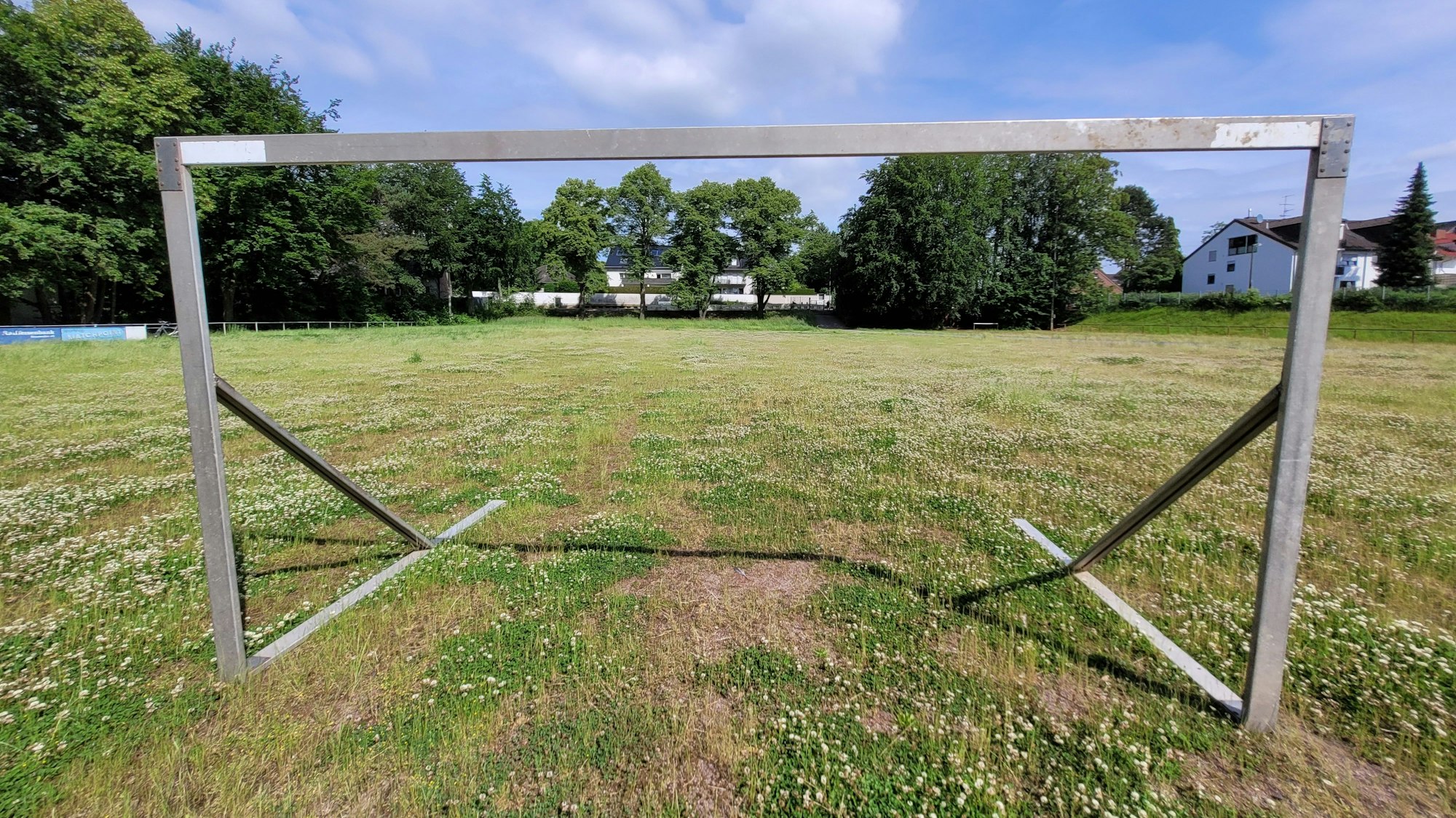 Ein Fußballtor ohne Netz steht auf einem von kleinwüchsigen Pflanzen überdeckten Aschenplatz. Im Hintergrund sind Häuser zu sehen.