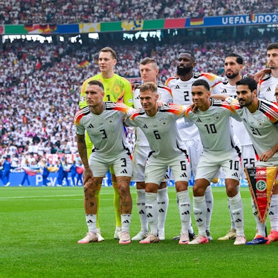 Die deutsche Nationalmannschaft posiert in zwei Reihen auf dem Spielfeld: Manuel Neuer, Toni Kroos, Antonio Rüdiger, Emre Can, Kai Havertz, Jonathan Tah, David Raum, Joshua Kimmich, Jamal Musiala, Ilkay Gündogan und Leroy Sané. Im Hintergrund sind voll besetzte Stadionränge zu sehen.